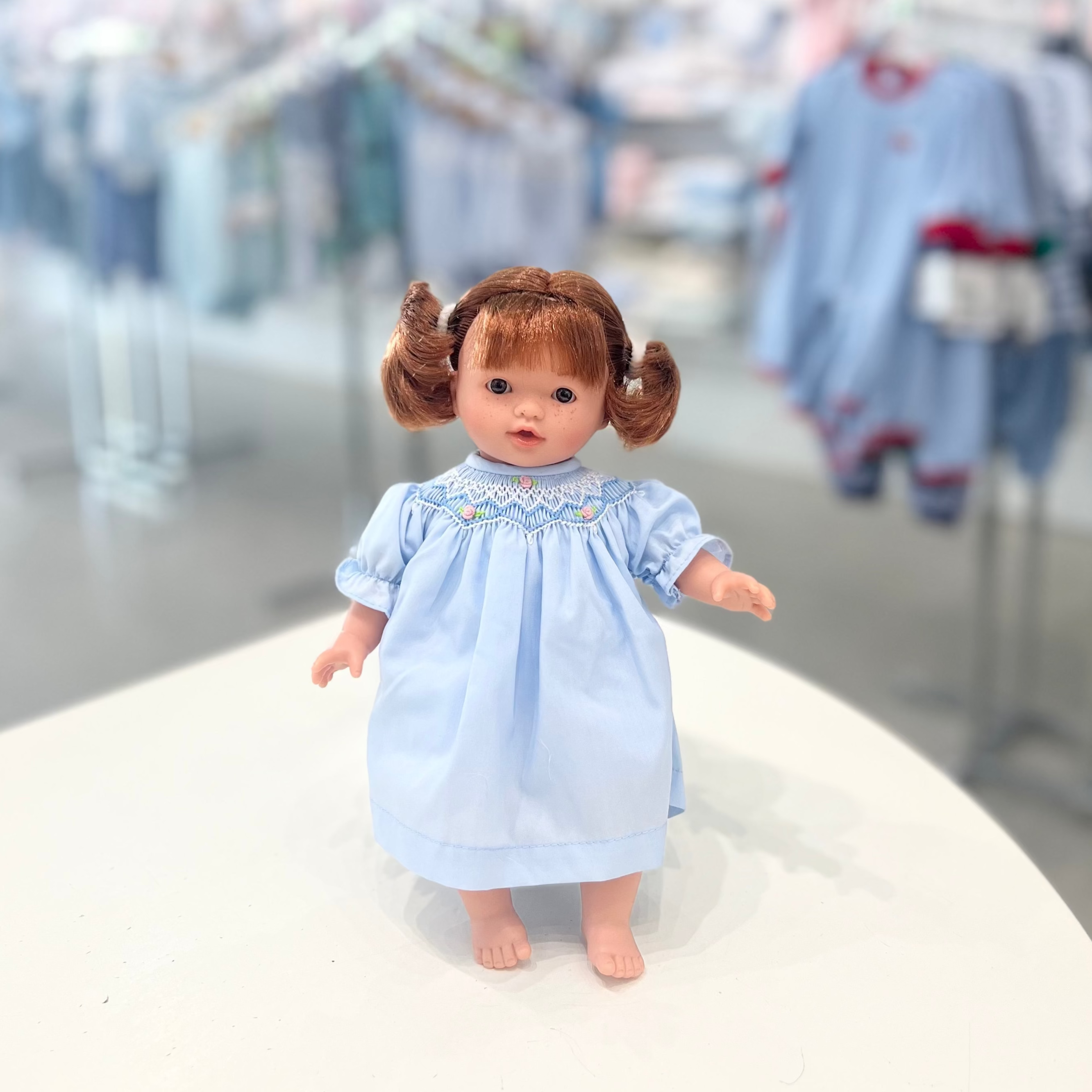 A 10-inch doll with red hair and brown eyes, dressed in a blue outfit with white trim, standing against a blurred background of similar dolls and accessories.